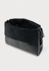 Mustast nahast ja seemisnahast õlakott, millel on volditav klapp ja nähtav seesmine lukuga tasku, valgel taustal.