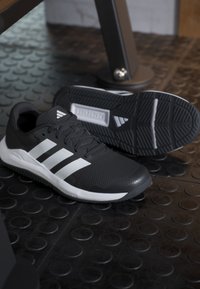 Chaussures de sport Adidas noires et blanches sur un sol de gym texturé noir près du matériel d'entraînement.