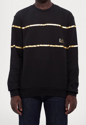 Homme portant une chemise noire à manches longues avec deux bandes horizontales dorées et un petit logo doré EA7 Emporio Armani sur la poitrine.