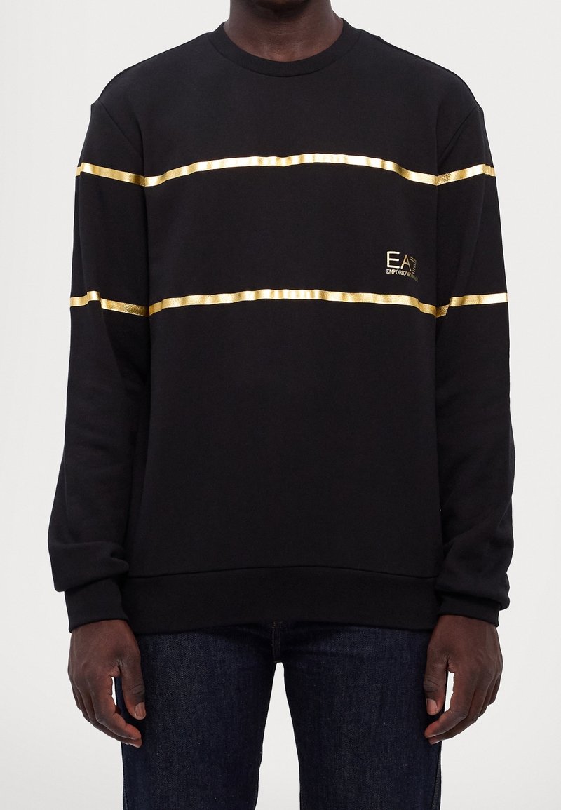 Homme portant une chemise noire à manches longues avec deux bandes horizontales dorées et un petit logo doré EA7 Emporio Armani sur la poitrine.
