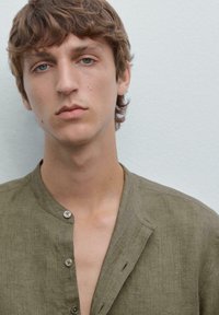 Young man with short, wavy hair and blue eyes, wearing a green buttoned shirt, gazes directly at the camera against a light gray background.