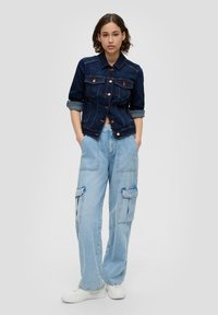 Donkerdenim jack met knoopsluiting, opgestikte zakken, opgerolde mouwen. Lichtblauwe cargobroek met meerdere zakken, casual pasvorm. Witte sneakers.