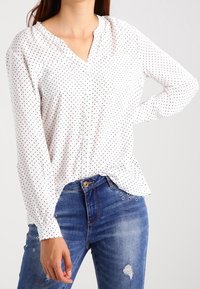 Blusa bianca a maniche lunghe con piccoli pois neri, presenta una scollatura a V e una vestibilità morbida, abbinata a jeans blu aderenti con decorazioni.