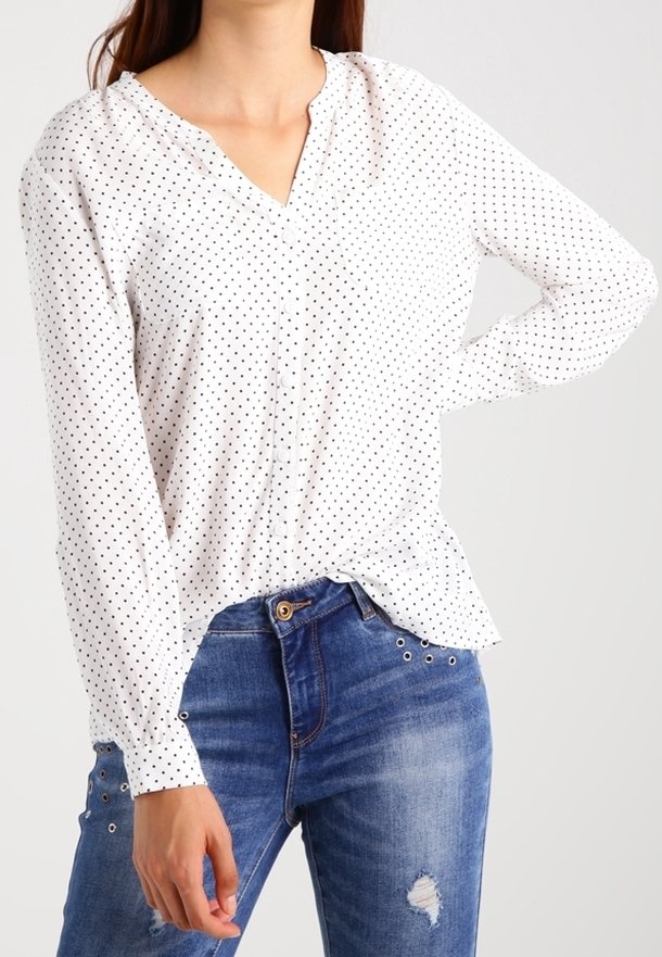 Blusa bianca a maniche lunghe con piccoli pois neri, presenta una scollatura a V e una vestibilità morbida, abbinata a jeans blu aderenti con decorazioni.
