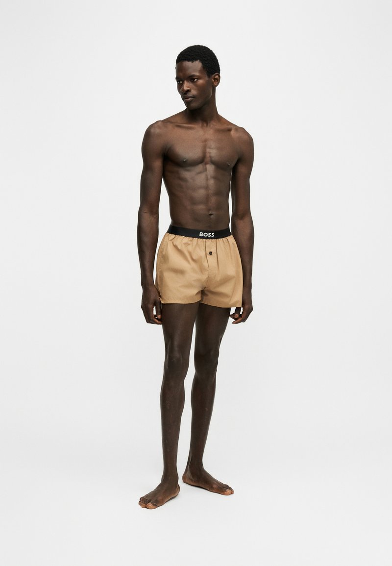 Homme mince à la peau foncée debout, pieds nus, portant un boxer beige avec une ceinture noire portant l'inscription BOSS, sur un fond blanc uni.