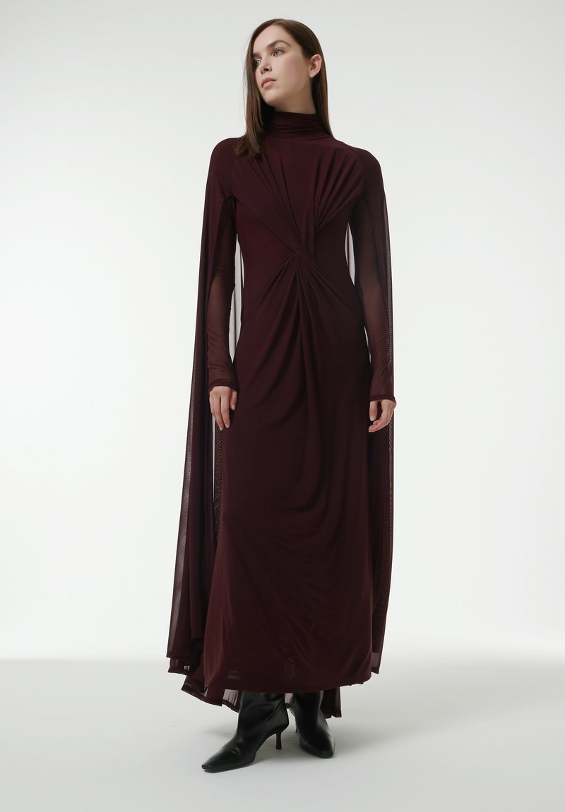 Burgunderfarbenes langes Kleid mit hohem Ausschnitt, drapiertem Oberteil und transparenten langen Ärmeln. Der Stoff weist aufwendige Faltungen und eine glatte Textur auf.