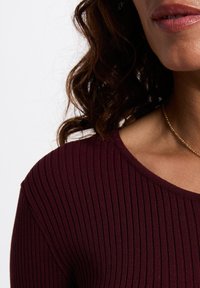 Top burdeos de canalé con escote redondeado; presenta un diseño ajustado y una textura fina. Collar dorado que acentúa el escote.