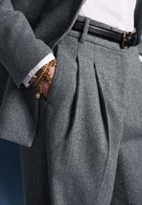Gray tailored suit with a textured wool blend, featuring pleated trousers, a black leather belt, and layered gold bracelets on a wrist.