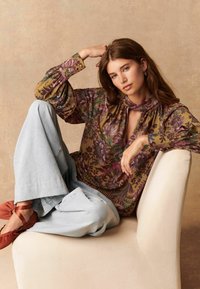 Blusa floreale a maniche lunghe in verde e viola su sfondo marrone, con scollatura a chiave; abbinata a jeans a gamba larga azzurri e ballerine rosse.