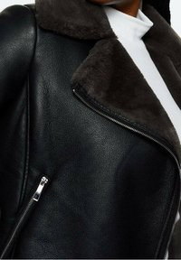 Veste en cuir noir avec un col en fausse fourrure, dotée d'une fermeture éclair argentée et d'une finition texturée. Le col contraste en gris foncé.