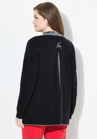 Femme en cardigan noir avec bordure grise et motif de renne dans le dos, associée à un pantalon rouge, se tenant contre un mur blanc.