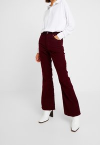 Pantalon évasé en velours côtelé bordeaux avec poches avant et fermeture par bouton, associé à une chemise blanche à col et manches longues, ainsi qu'à des bottes blanches à talons.
