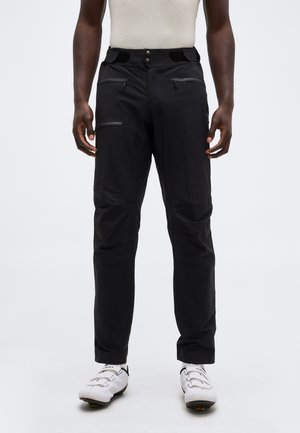 Vyras juodomis lauko kelnėmis su užtrauktuku ir baltais dviračių batais, stovintis priešais vienodus fono paviršius.