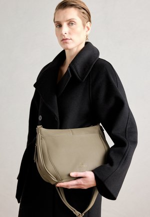 Vrouw in een zwarte jas die een taupekleurige lederen schoudertas met verstelbare riem vasthoudt tegen een neutrale achtergrond.