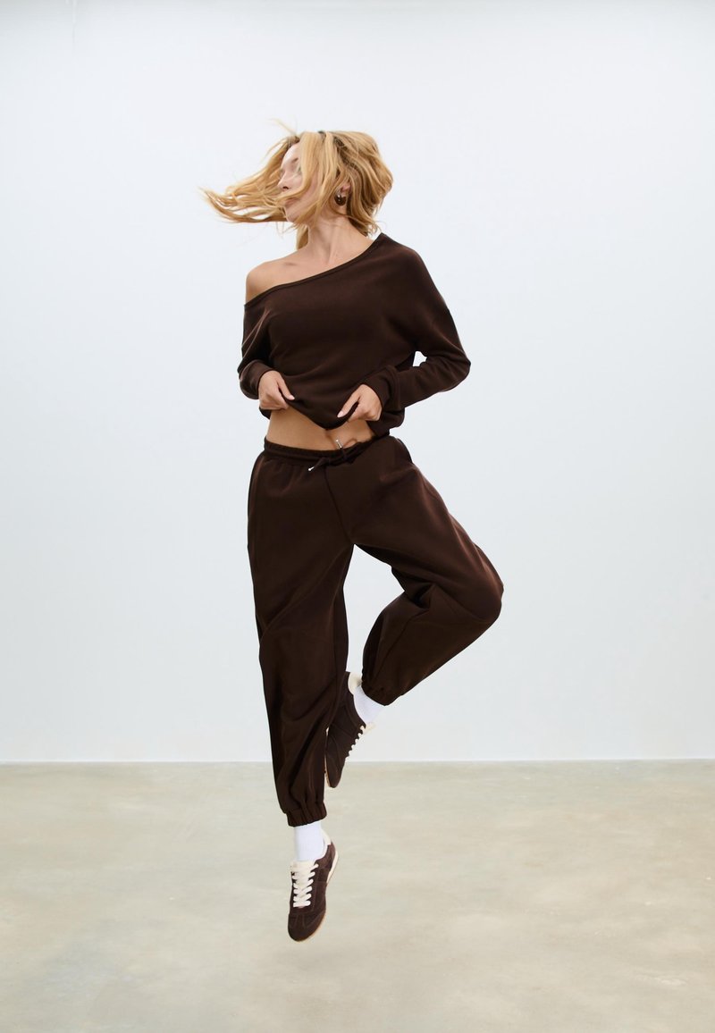 Blondynka skacząca w powietrzu, ubrana w ciemnobrązową bluzkę odsłaniającą ramiona, pasujące spodnie dresowe, białe skarpetki i ciemne tenisówki na jednolitym tle.