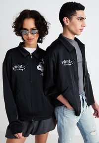 Črne jakne z zadrgo, širok kroj in vezeni detajli. Model je kombiniral s sivo majico in kavbojkami, opremljen z sončnimi očali.