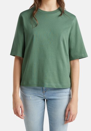 Kvinde iført en ensfarvet grøn kortærmet skjorte og lyseblå jeans, vist fra skuldre til hofter mod en hvid baggrund.