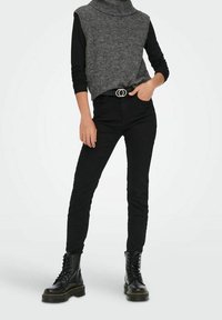 Personne portant un pull à col roulé sans manches gris sur une chemise noire à manches longues, un pantalon noir, une ceinture noire et des bottes noires à lacets.