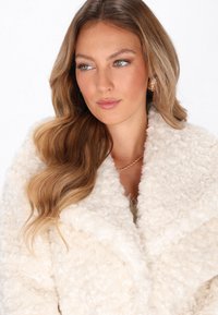 Vrouw met lang golvend blond haar, gekleed in een gestructureerde witte bontjas, gouden kettinghalsketting en parel oorbellen, die iets naar de zijkant kijkt.
