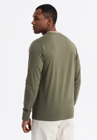 Uomo che indossa una camicia verde oliva a maniche lunghe e pantaloni bianchi, in piedi con la schiena rivolta in avanti, guardando a sinistra.