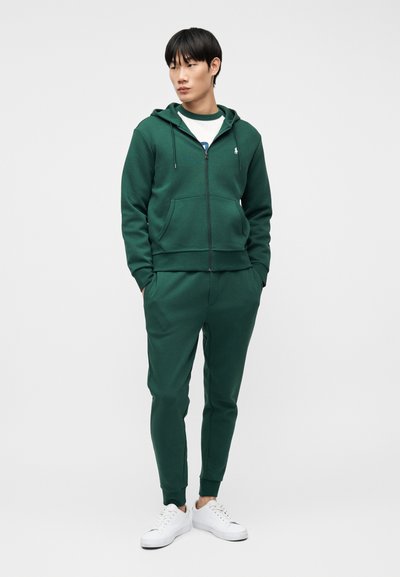 Άνδρας που φοράει σκούρο πράσινο φουλάρι με φερμουάρ και ταιριαστό παντελόνι φόρμας, με λευκά αθλητικά παπούτσια, στέκεται με τα χέρια στις τσέπες μπροστά σε απλό φόντο.