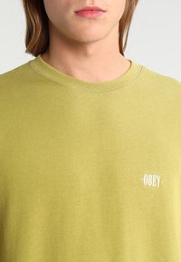 Oliveně zelený bavlněný svetr s kulatým výstřihem. Obsahuje malé bílé logo "OBEY" na hrudi a viditelné švy.