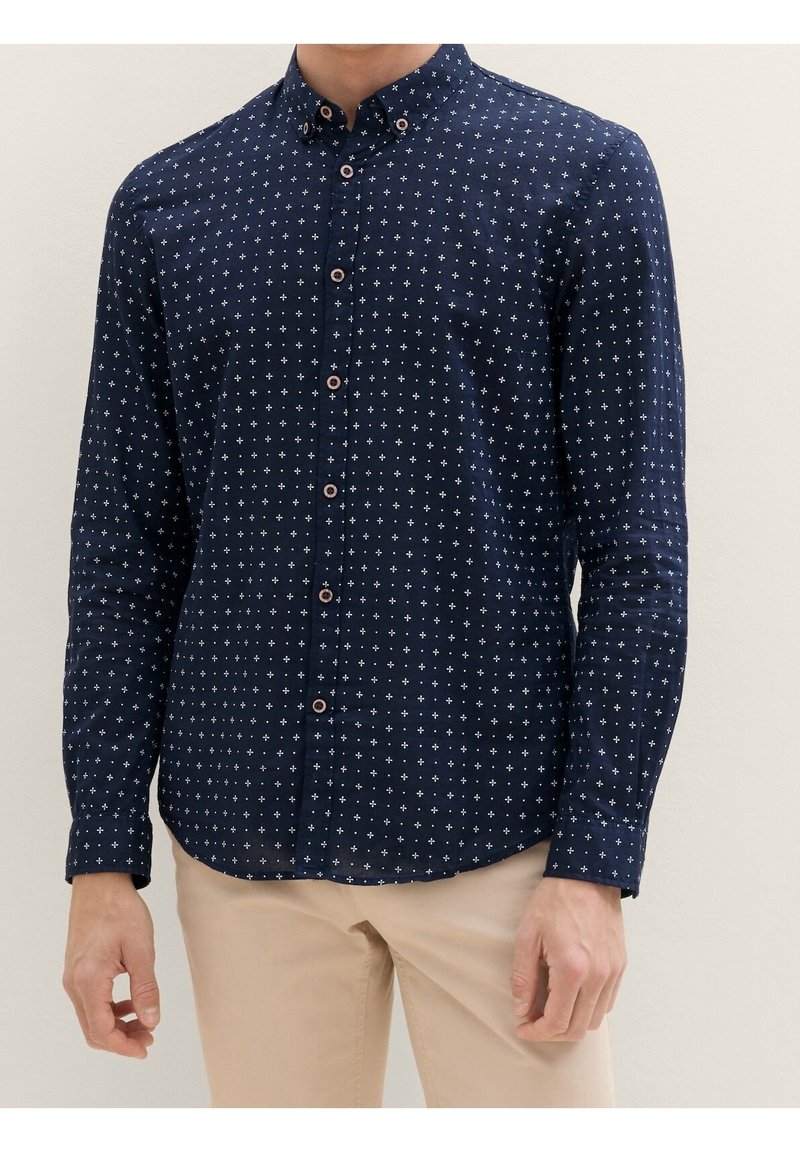 Homme portant une chemise bleu marine à boutons avec des motifs de croix blanches et un pantalon beige, debout devant un fond clair uni.
