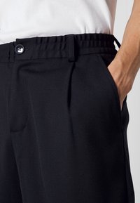 Schwarze Hose aus strukturiertem Material, mit einem elastischen Bund und einem Knopfverschluss. Seitentaschen und gefältelte Details sind sichtbar.