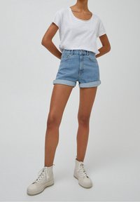 Person som bär uppvikta ljusblå denimshorts, vit t-shirt med rund halsringning och vita snörskor med hög skaft, står mot en enfärgad bakgrund.