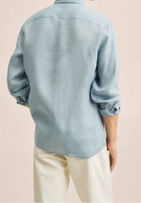 Jasnoniebieska lniana koszula z długimi rękawami podwiniętymi, o subtelnej fakturze i zaokrąglonym dole, noszona z jasnymi spodniami.