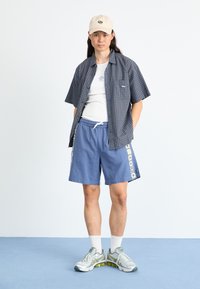 Kortsleeve rutete skjorte i marineblå og grønn, hvit tank topp, blå shorts med dekorativ sidepanel, og sølvfargede joggesko.