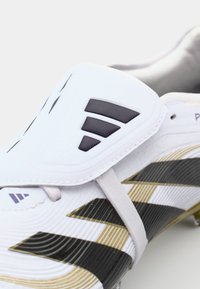Baltie futbolsneakers ar melniem un zeltainiem akcentu svītrojumiem. Ir strukturēts augšdaļa un aerodinamisks dizains. Iekļauj atšķirīgu logo elementu.