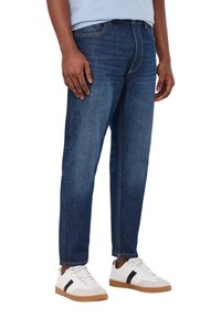 Jeans in denim blu scuro con un design a gamba dritta, caratterizzati da cuciture più scure. Abbinati a sneakers bianche con dettagli neri.