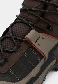 Καφέ πεζοπορικό παπούτσι με υφασμάτινο επάνω μέρος με υφή, ενισχυμένες συνθετικές επικάλυψεις, μαύρα κορδόνια και κόκκινες λεπτομέρειες στο πλάι.