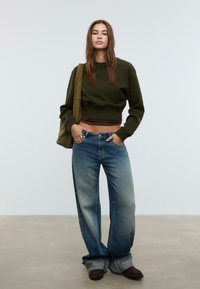 Donna che indossa un maglione corto verde oliva, jeans blu sbiaditi a gamba larga, scarpe marroni, mentre porta una borsa a tracolla verde oliva, in piedi contro uno sfondo semplice.