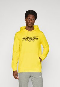 Gul hættetrøje med en frontlomme og justerbar snøre i hætten, med et sort Dortmund-logo over brystet. Grå sweatpants vist nedenfor.