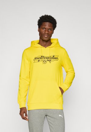 Gul hættetrøje med en frontlomme og justerbar snøre i hætten, med et sort Dortmund-logo over brystet. Grå sweatpants vist nedenfor.
