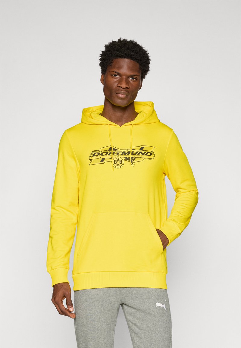 Gul hættetrøje med en frontlomme og justerbar snøre i hætten, med et sort Dortmund-logo over brystet. Grå sweatpants vist nedenfor.