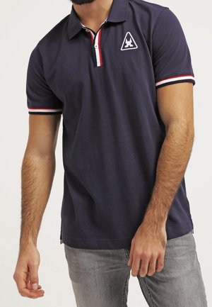 Mann trägt ein marineblaues Poloshirt mit rot-weiß-blau gestreiftem Kragen und Ärmelbündchen, kombiniert mit hellgrauen Jeans.