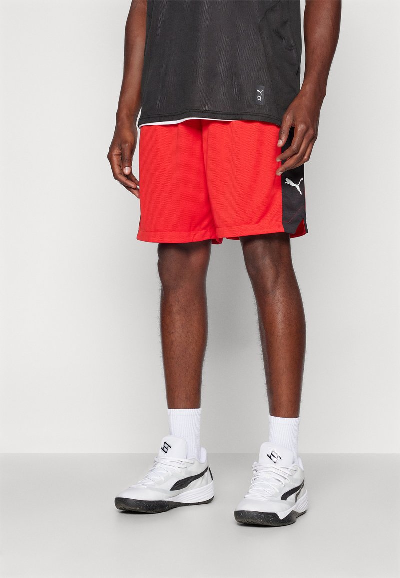 Røde treningsshorts med mesh-textur, med et svart sidepanel med logo. Kombinert med hvite joggesko og hvite sokker.