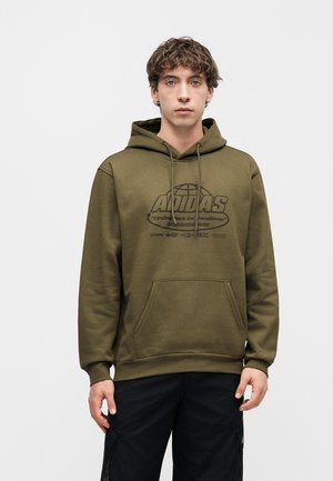 Młody mężczyzna w oliwkowo-zielonej bluzie z kapturem Adidas z czarnym tekstem i grafiką, stojący na jednolitym, jasnoszarym tle.