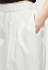 Main de femme dans la poche latérale d'une jupe plissée blanche taille haute, avec une large ceinture et une légère fente.