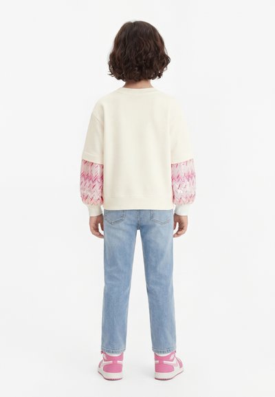 Παιδί με κοντά σγουρά μαλλιά στέκεται με την πλάτη, φοράει κρεμ φούτερ με ροζ μανίκια με σχέδια, ανοιχτό μπλε τζιν και ροζ αθλητικά παπούτσια.