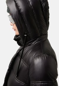 Osoba ubrana w błyszczącą czarną kurtkę puchową z podniesionym kapturem, zieloną dzianinową czapkę i dużą lustrzankę na białym tle.