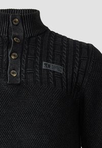 Schwarzer Strickpullover mit hohem Rippkragen, drei goldfarbenen Knöpfen, Zopfmuster an den Schultern und einem Stoff-Logoetikett.
