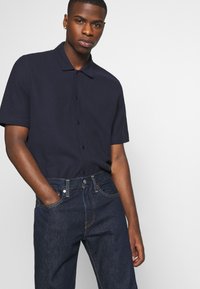 Polo de manga corta en color navy con cierre de botones, combinado con jeans de mezclilla oscuros. El polo cuenta con un cuello clásico y una textura lisa.