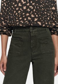 Pantalons en denim vert olive taille haute, avec poches latérales et couture contrastée ; portés avec un blouse marron à motifs.