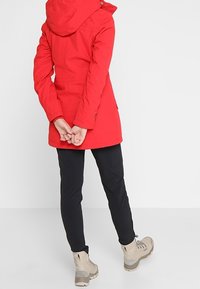 Parka aislante roja con capucha, puños elásticos y bolsillos con cremallera, combinada con pantalones negros y botas de senderismo beige.