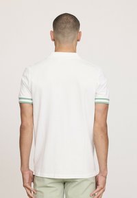 Polo shirt blanc avec des poignets à rayures vertes ; manches courtes ; col simple ; texture lisse ; vue de dos, porté avec un pantalon vert clair.