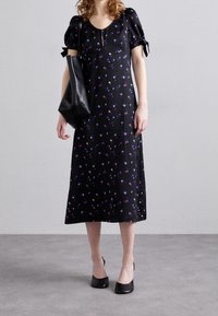 Kvinna klädd i en svart midiklänning med lila blommönster, puffärmar med snörning, svarta slingbackklackar och bär en svart läderhandväska.